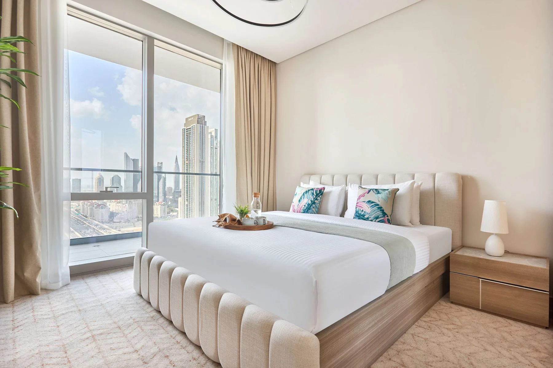 A bright bedroom with a large bed beside windows overlooking the city.
