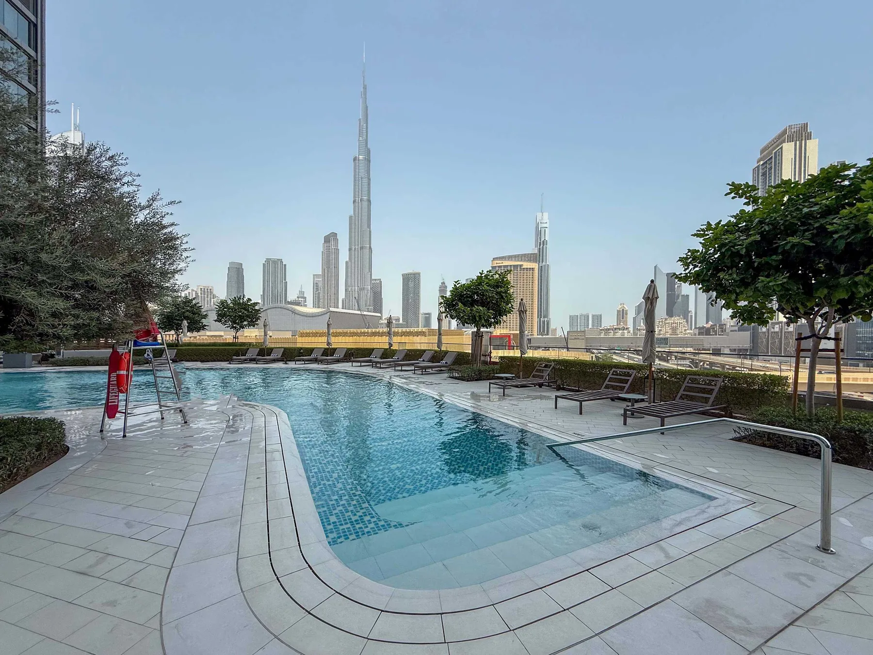 An outdoor swimming pool overlooks a city skyline, with lounge seating along the deck.