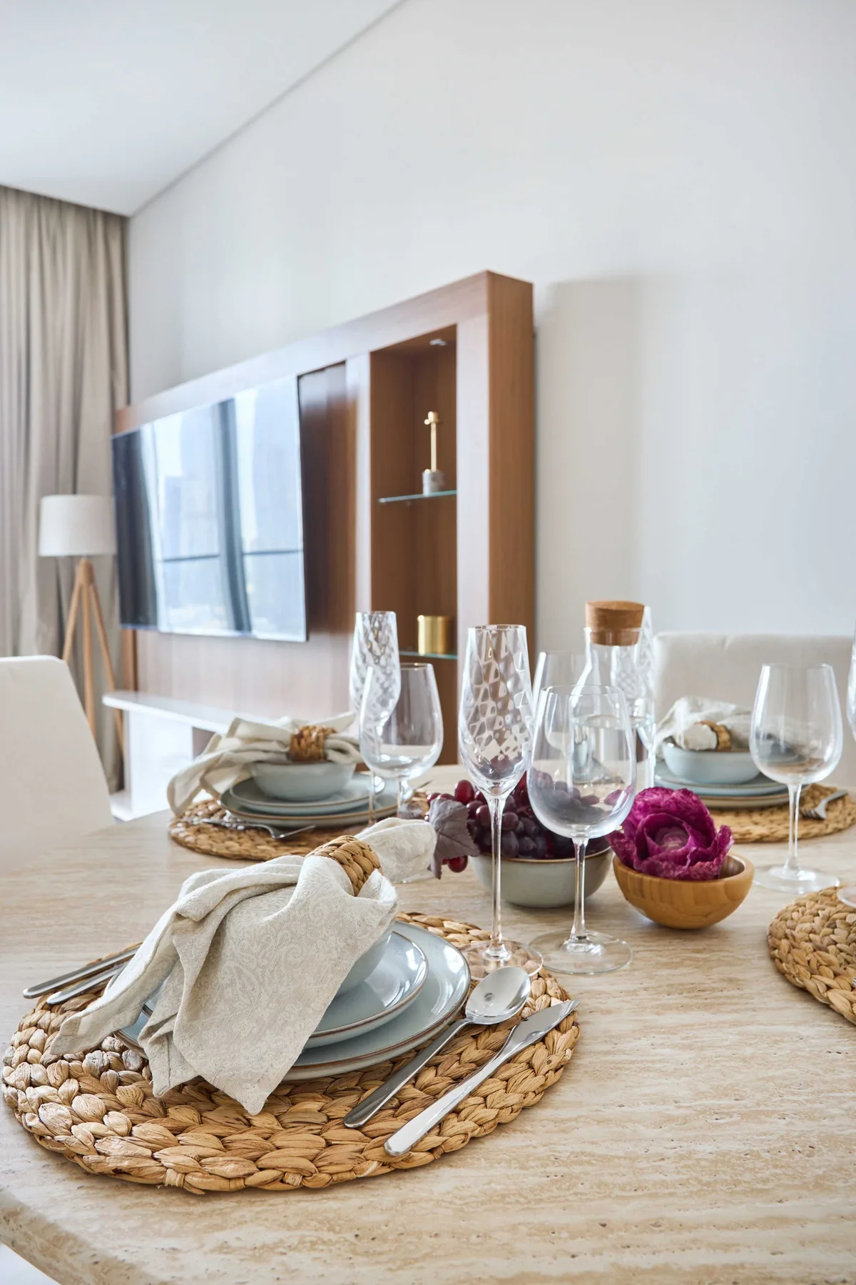 A dining table is set with plates, woven placemats, and glassware in a bright room.