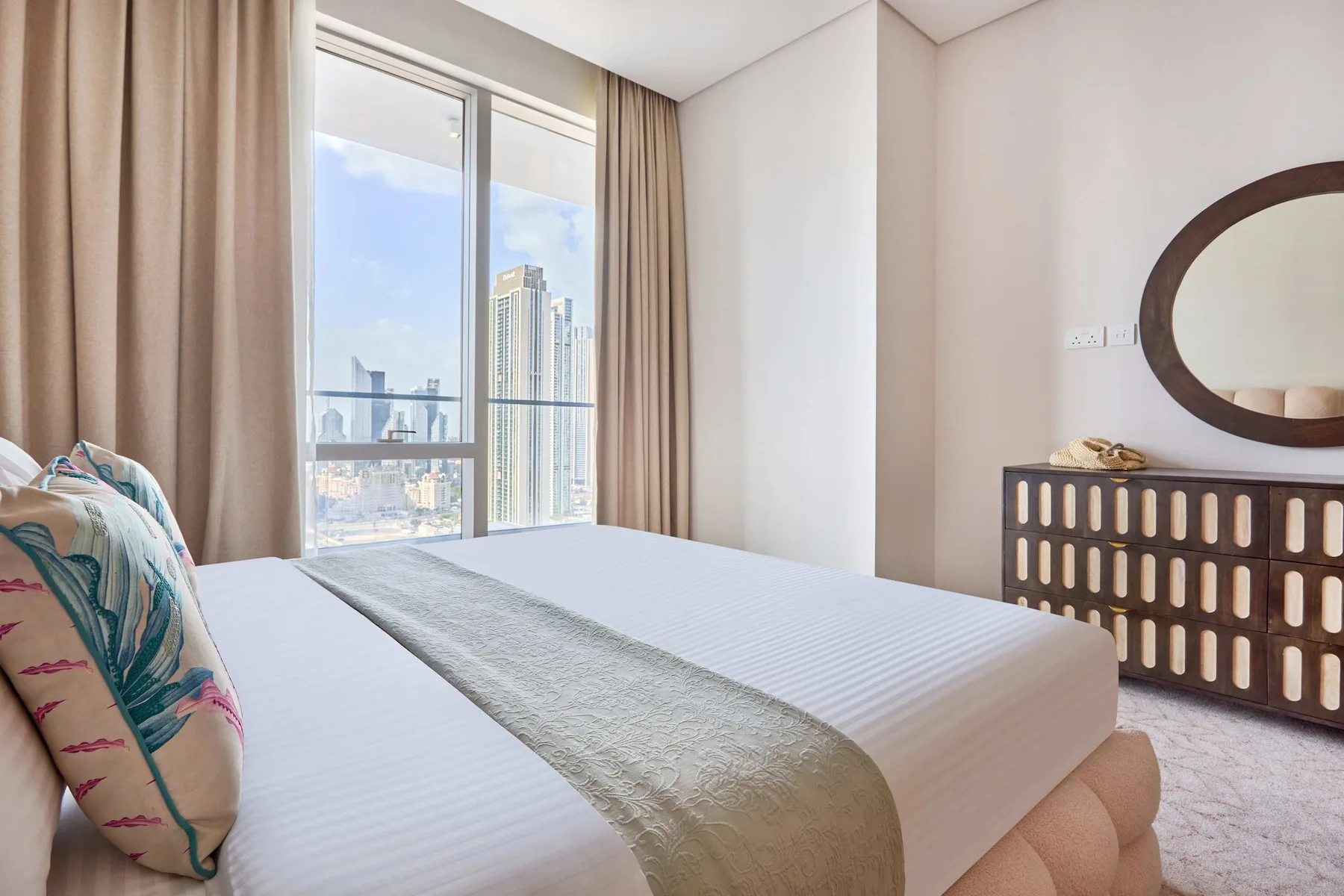 A bright bedroom with a bed beside a large window overlooking city buildings.