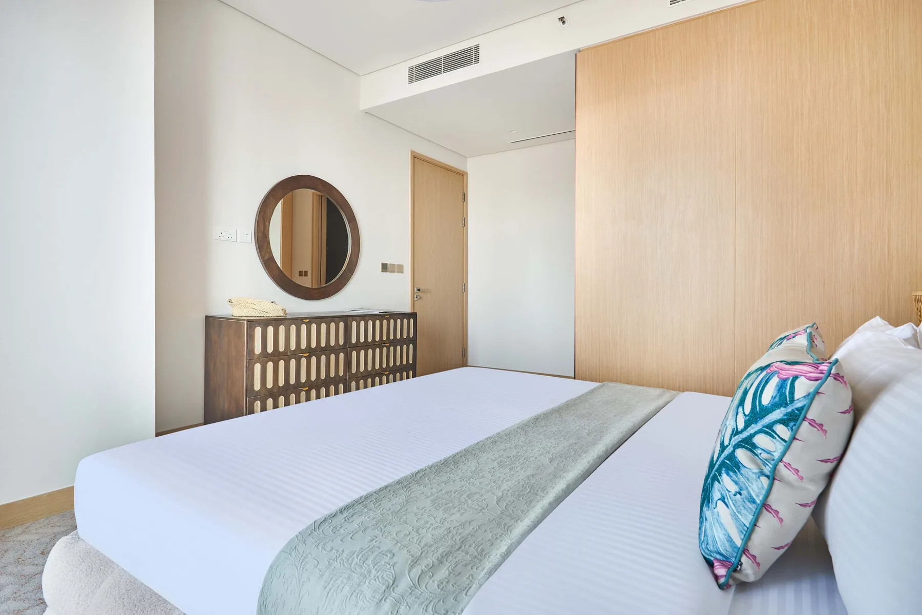 A bedroom with a neatly made bed, a decorative pillow, and a round wall mirror.