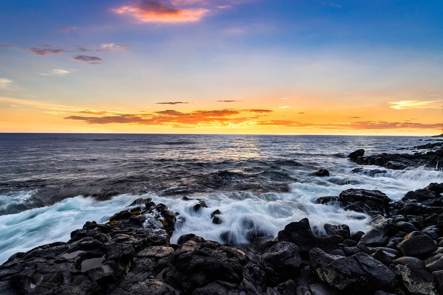 Atardecer sobre el océano, con olas bañando una costa rocosa.