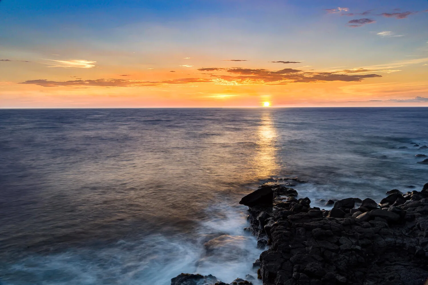 Atardecer sobre el océano, con el sol reflejado en el agua junto a una costa rocosa.