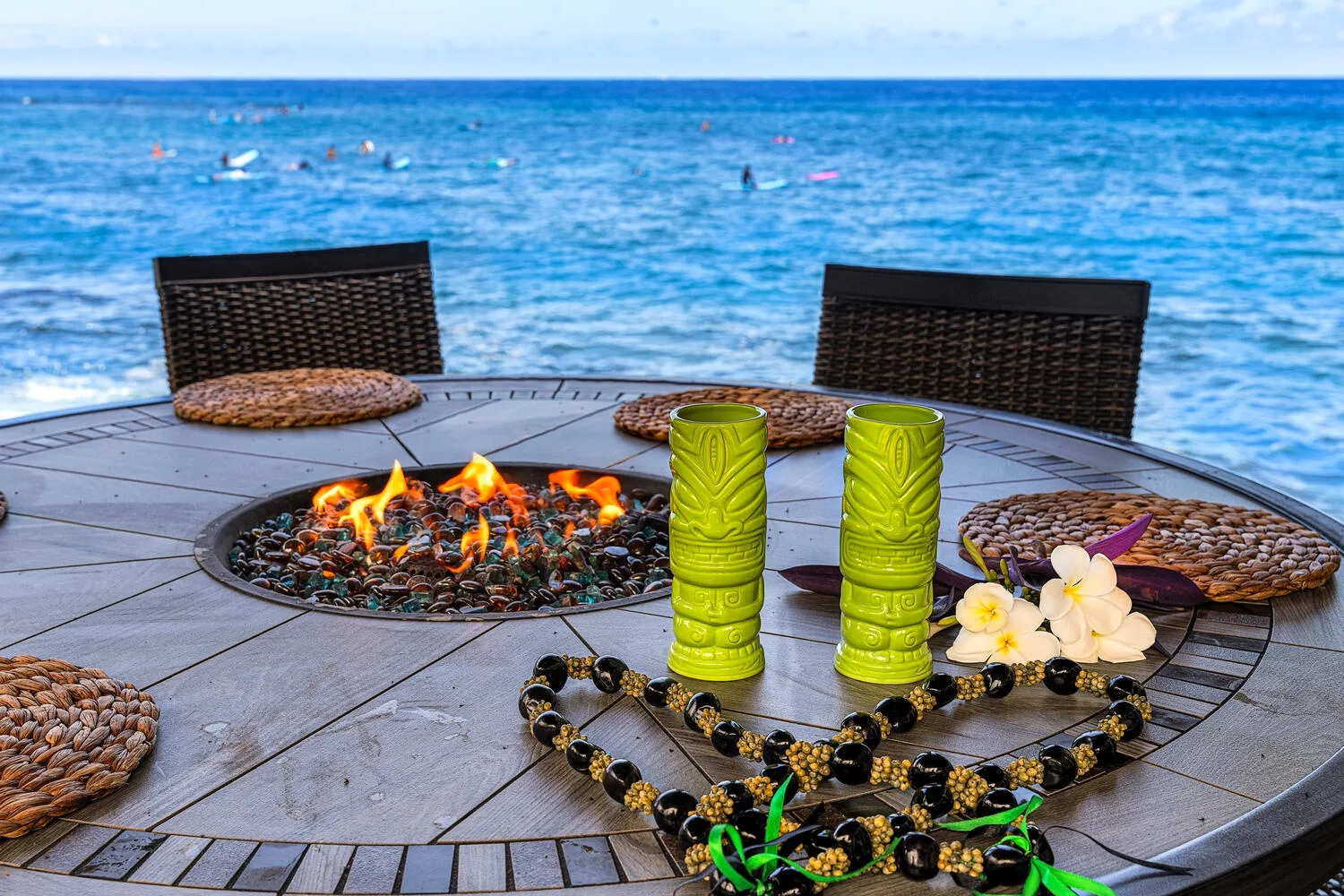 Una mesa en un patio junto al mar tiene un fogón encendido, vasos verdes y collares decorativos.
