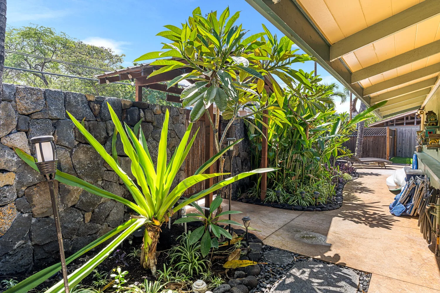 Un jardín tropical bordea un muro de piedra junto a un pasillo exterior cubierto.