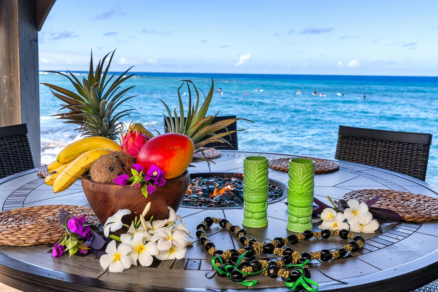 Una mesa de comedor junto al mar muestra frutas tropicales, flores y vasos verdes.