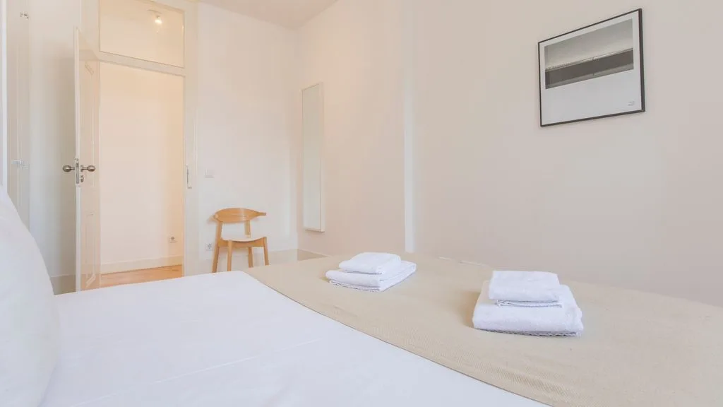 Un dormitorio luminoso con una cama, toallas dobladas y un pequeño asiento de madera.