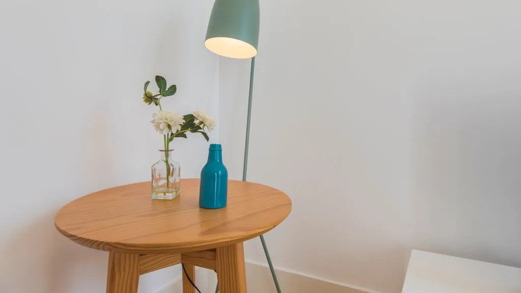 Una pequeña mesa redonda de madera está bajo una lámpara de pie verde, con flores y un jarrón azul.