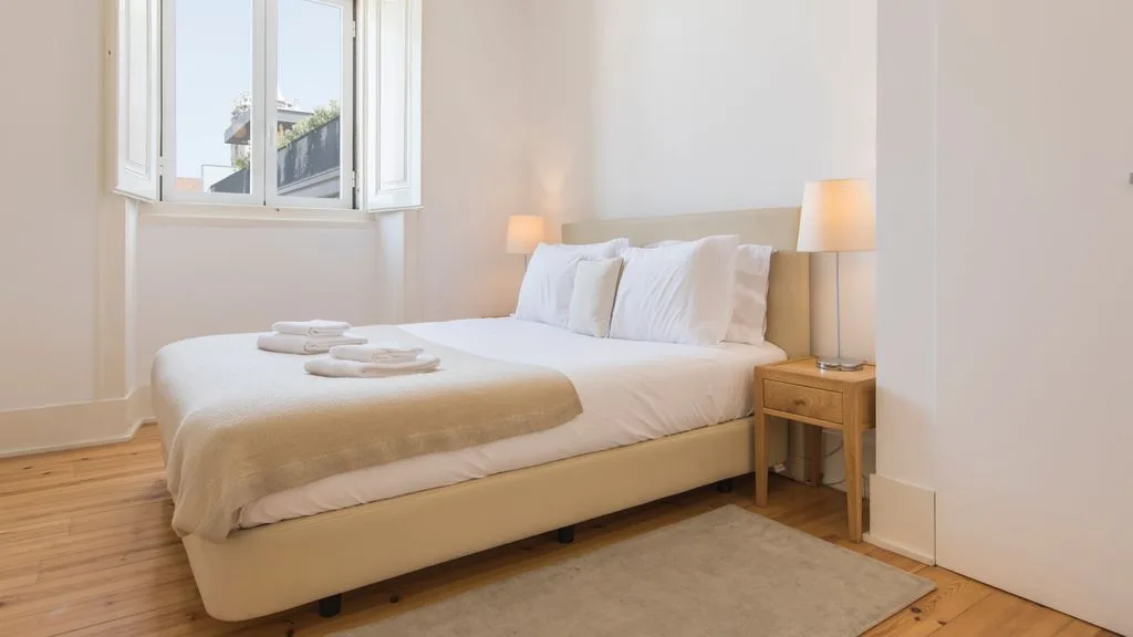 Un dormitorio luminoso con una cama bien hecha junto a una ventana abierta.