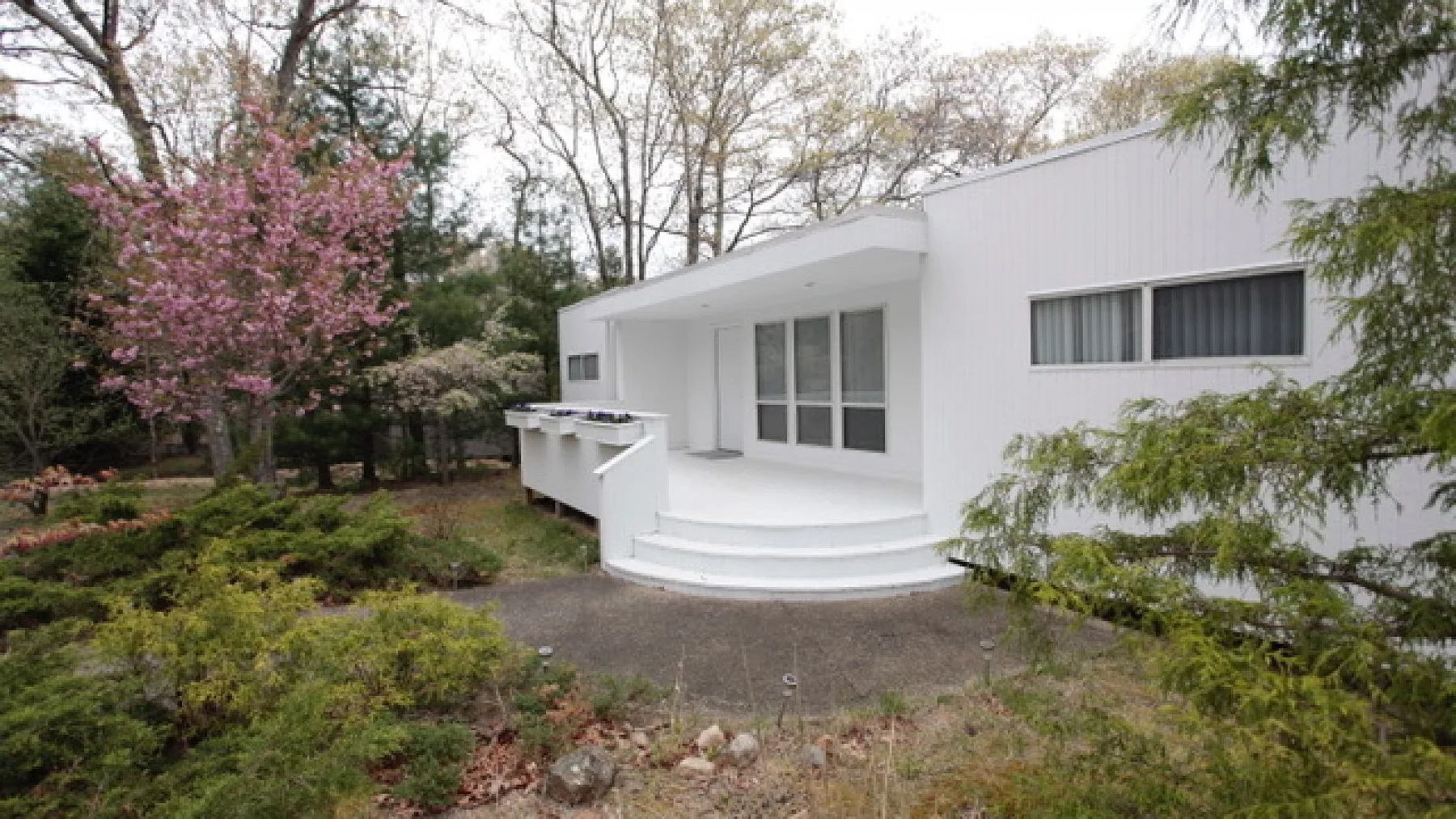 A white modern house opens onto a curved porch surrounded by trees and shrubs.