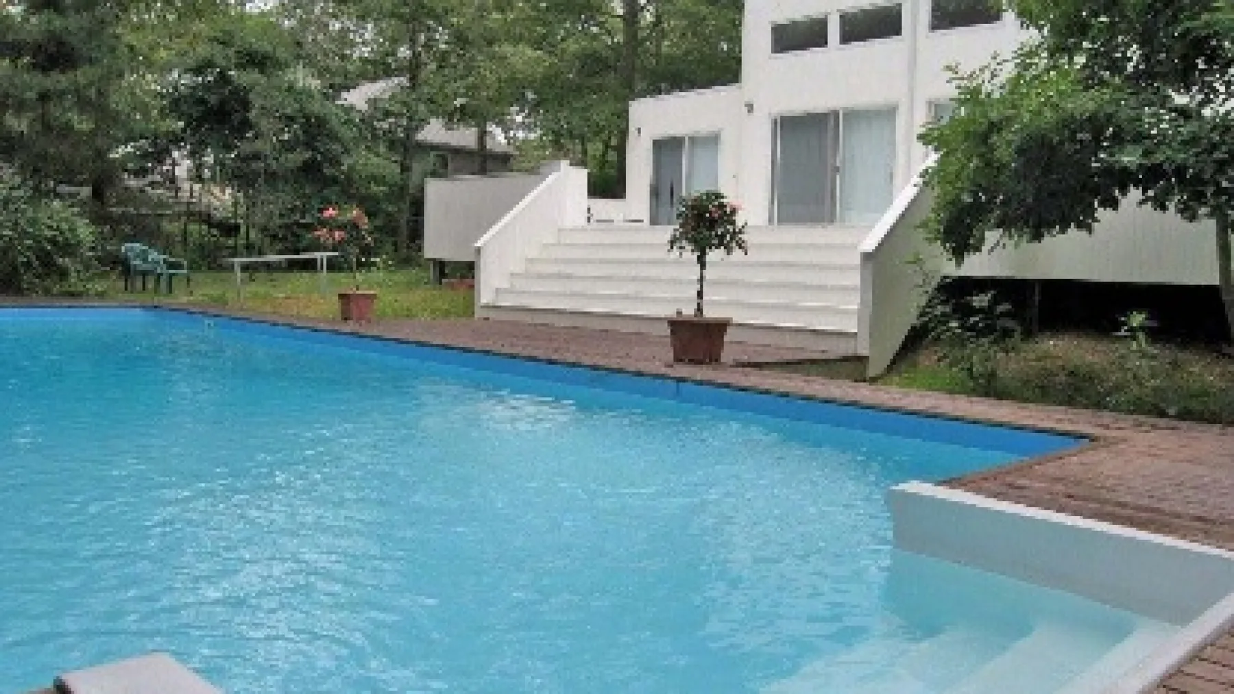 An outdoor swimming pool beside a white building and leafy trees.