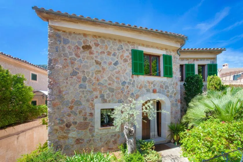 A stone house with green shutters sits among leafy garden plants.