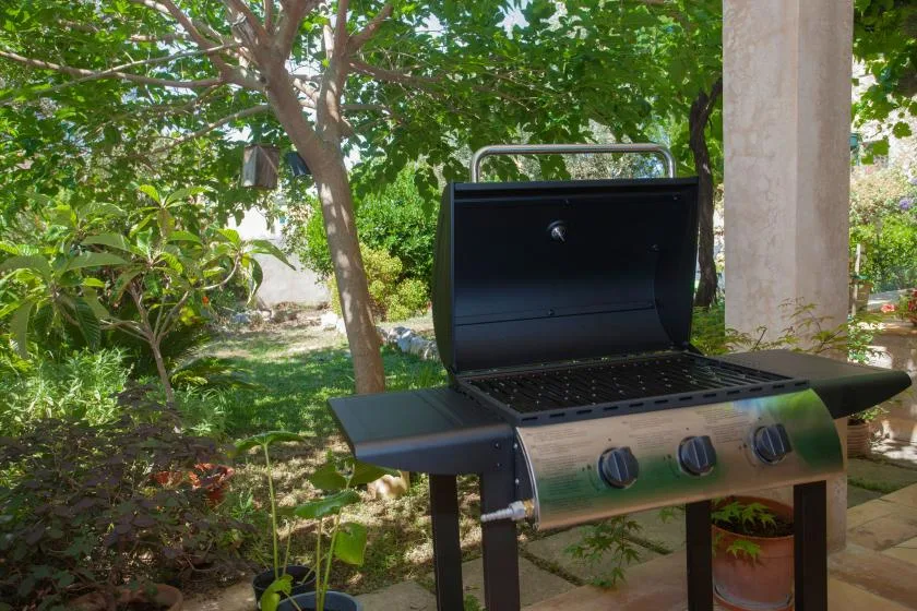 An outdoor grill stands beneath leafy trees in a garden.