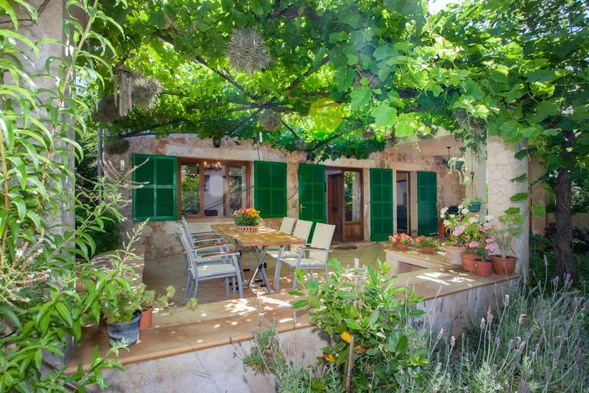 A leafy outdoor patio with a table, seating, and green-shuttered windows.