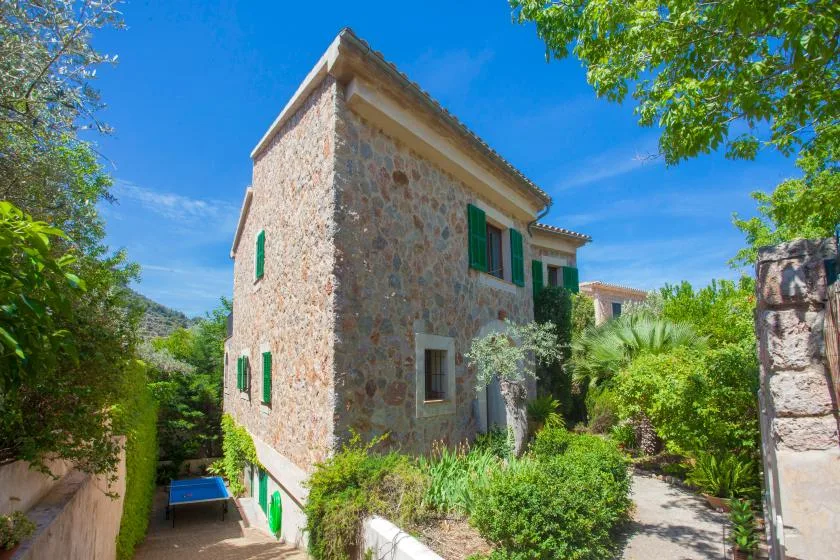 A stone house with green shutters stands among leafy garden plants.