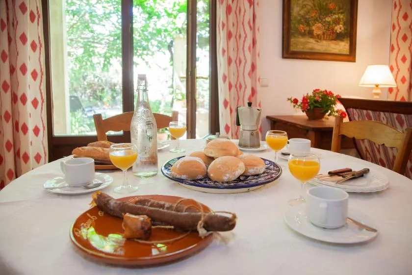 A dining table set with bread, drinks, and cups sits beside a bright window.