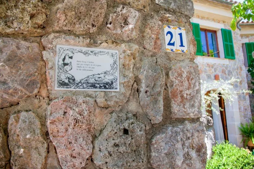 A stone exterior wall displays a small illustrated plaque and a blue number sign.