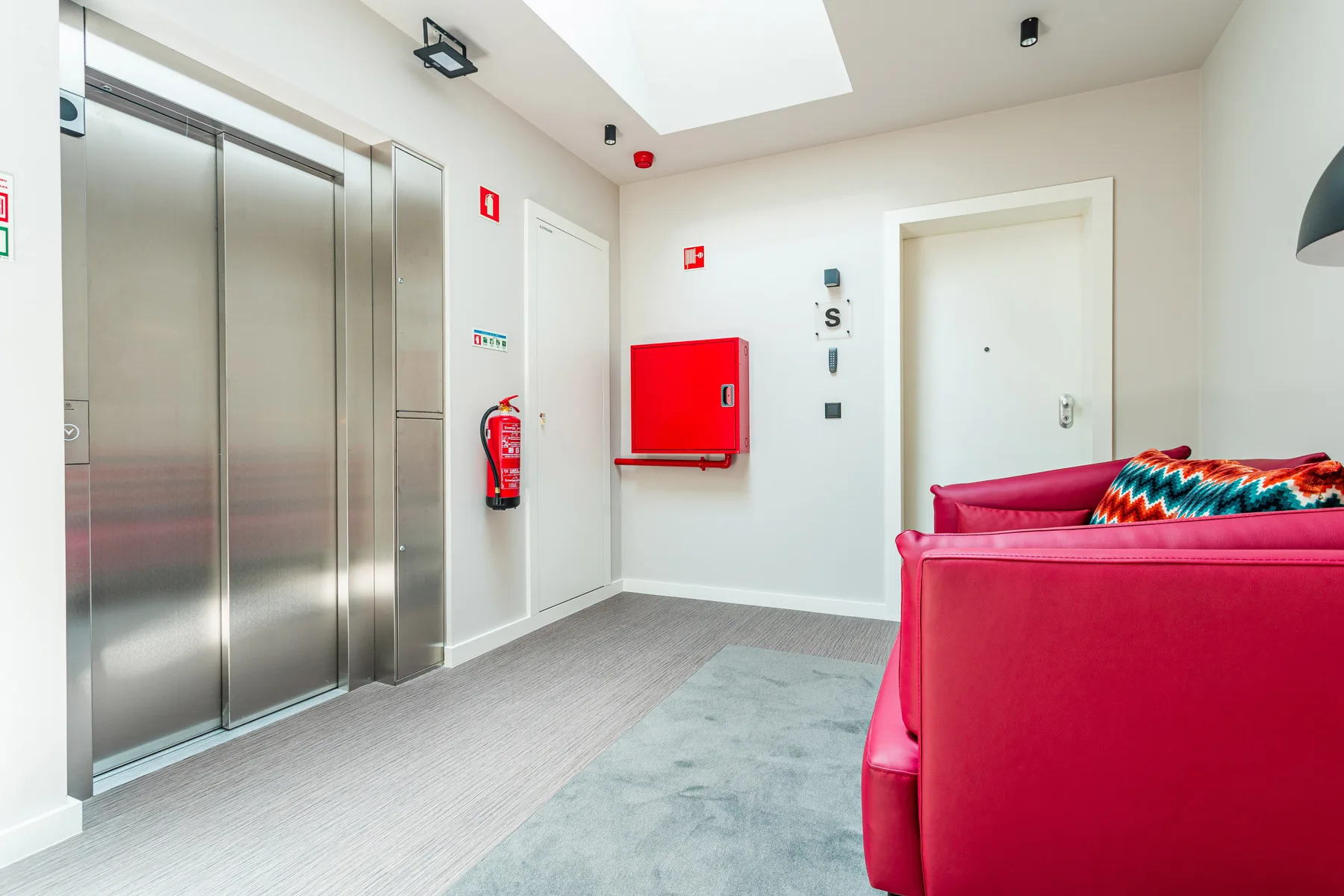 An apartment building lobby with elevator doors, a red wall cabinet, and seating.