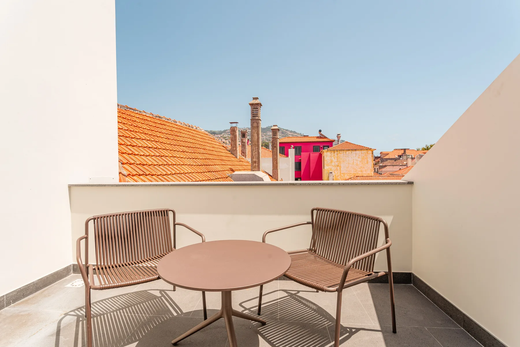 A sunny terrace with two seats and a round table overlooking nearby rooftops.