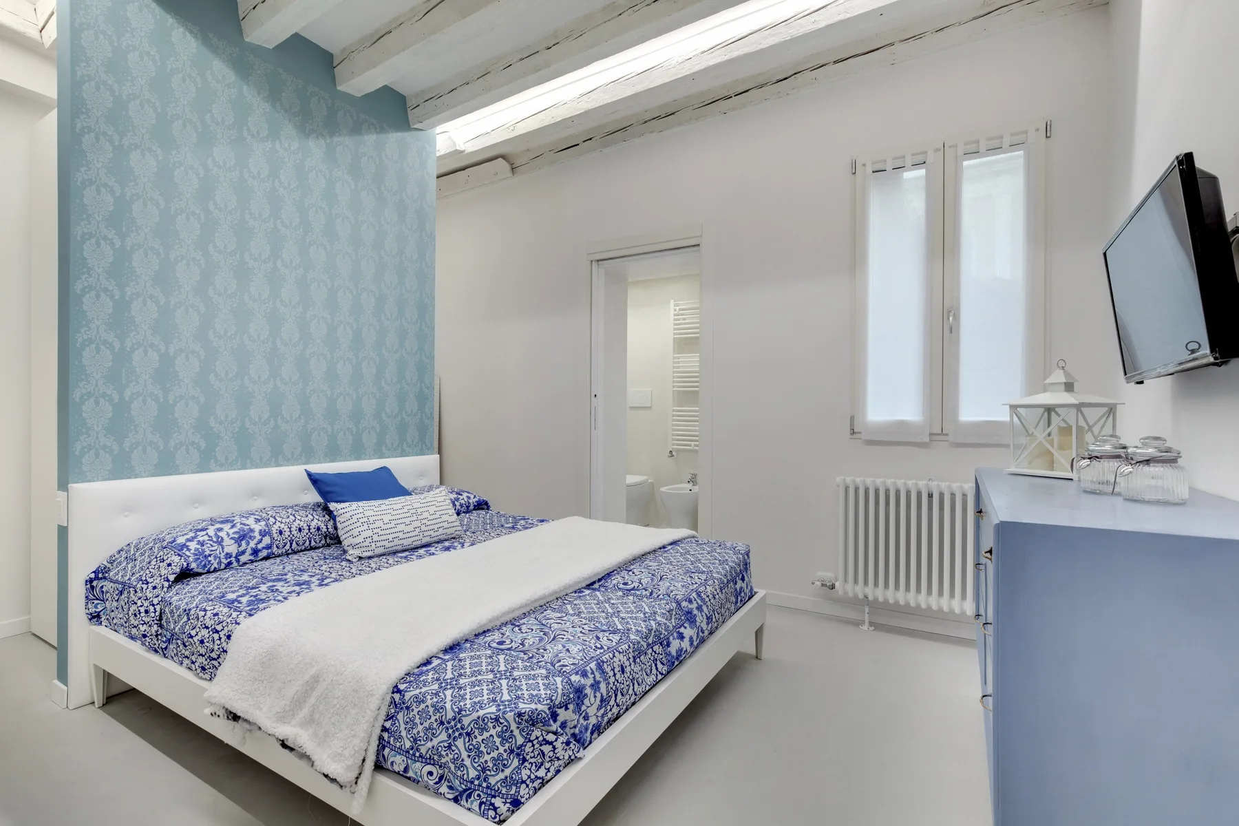 Un dormitorio con una colcha azul estampada y un televisor montado en la pared.