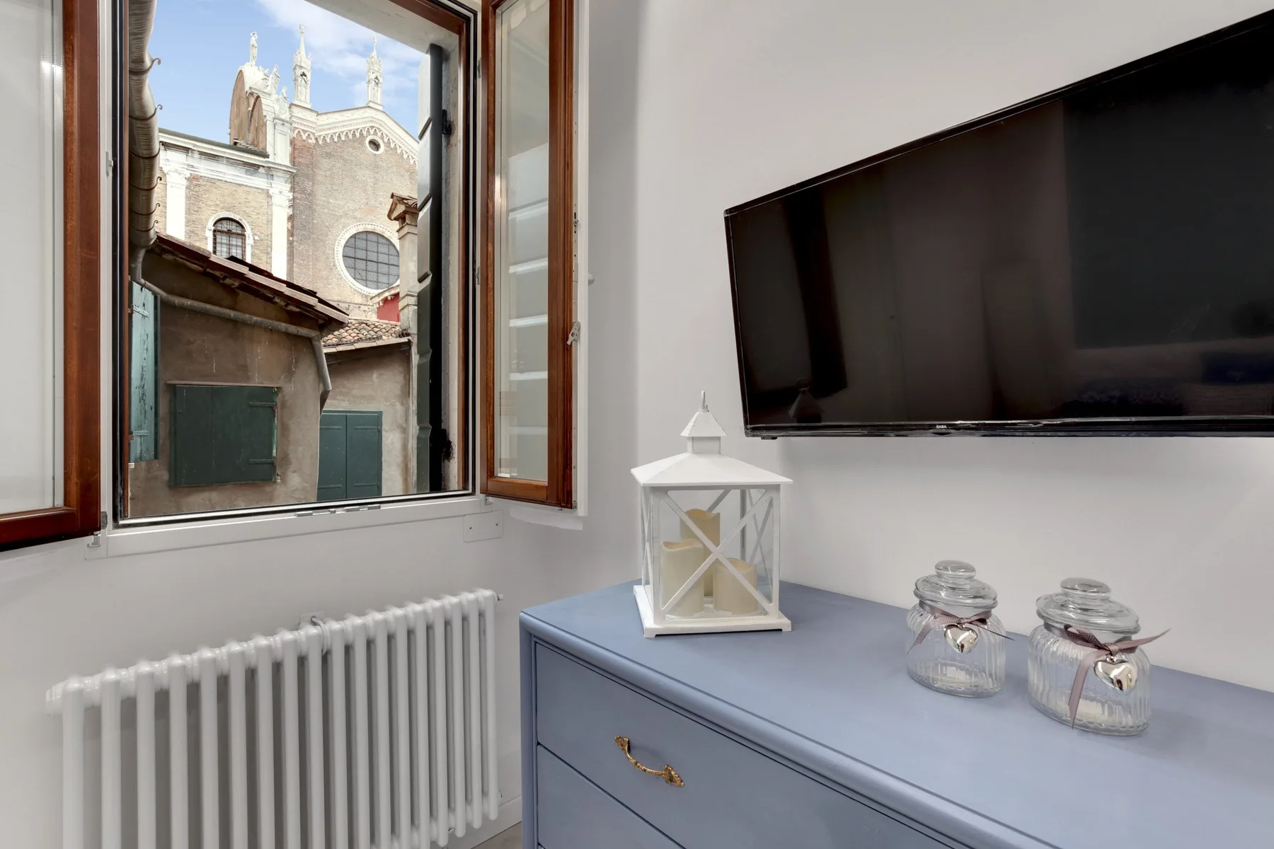 Una habitación con un televisor sobre una cómoda y una ventana abierta con vistas a un edificio.