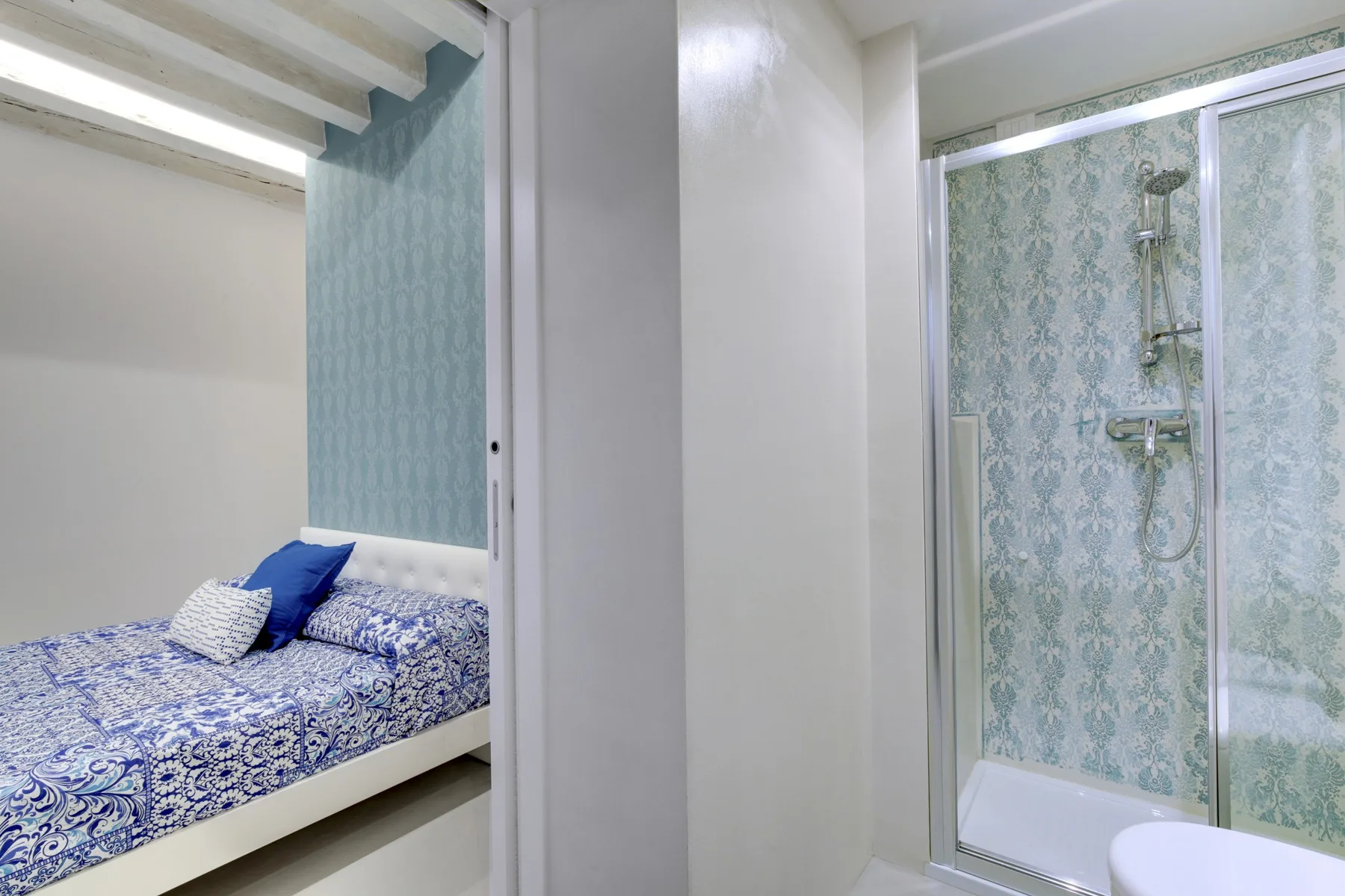 Un dormitorio con ropa de cama azul estampada junto a una ducha con mampara de vidrio.