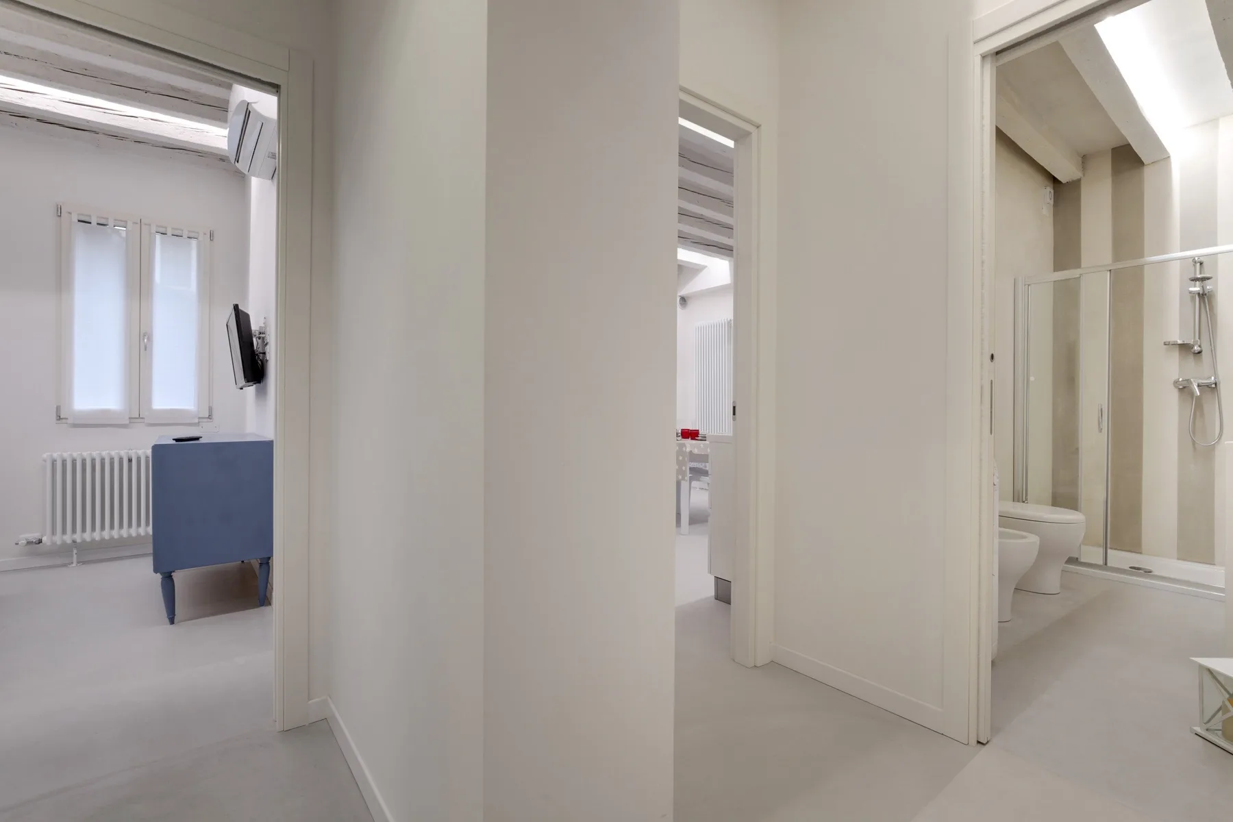 Un pasillo blanco da a un baño luminoso y a una habitación contigua.