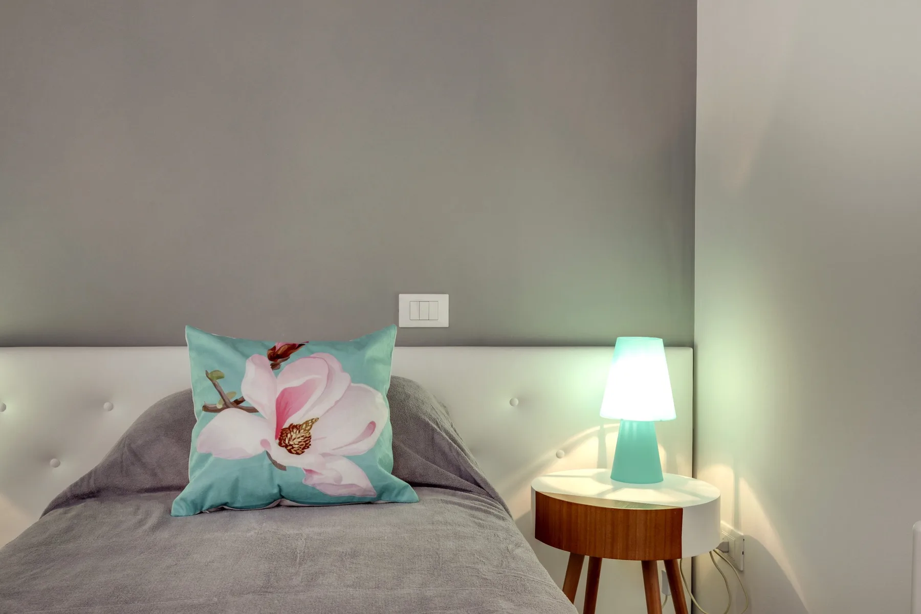 Un dormitorio con una cama, un cojín floral y una lámpara encendida.