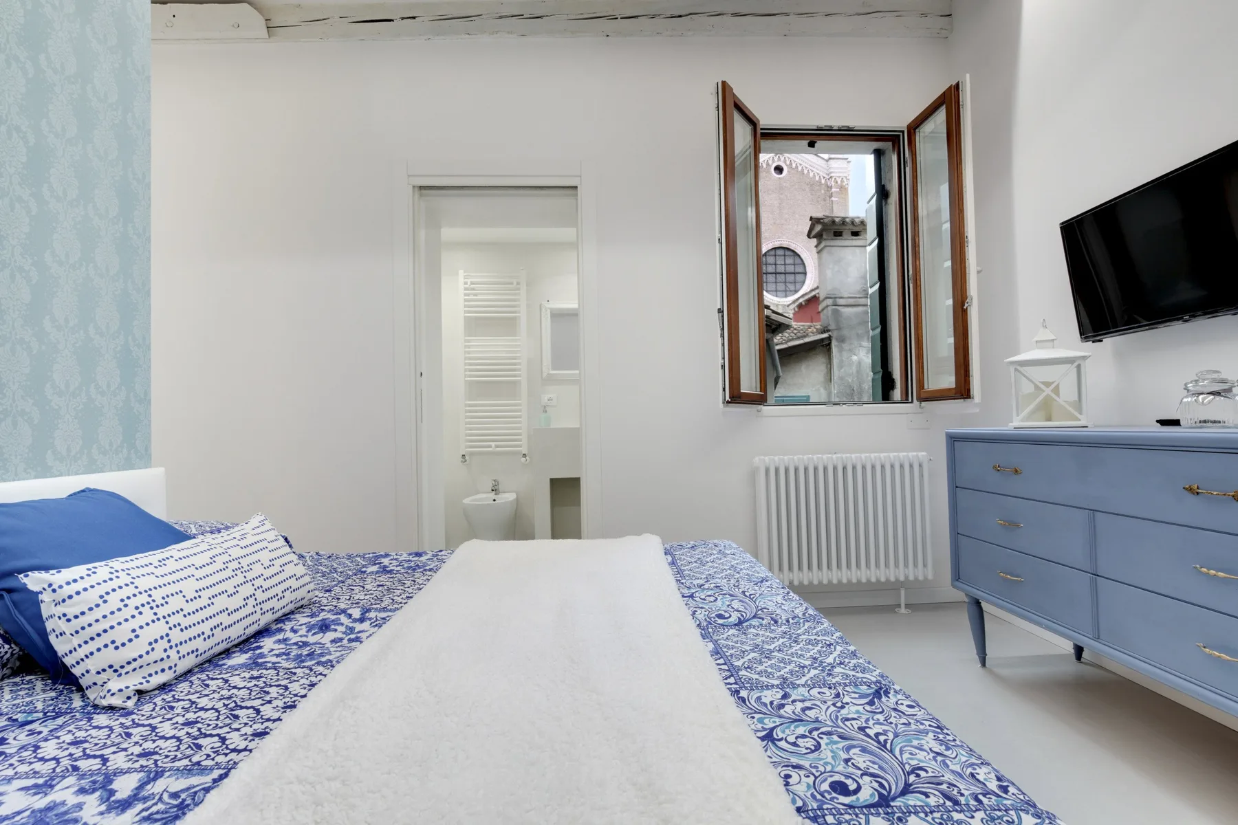 A bedroom with blue patterned bedding and a window beside a blue dresser.