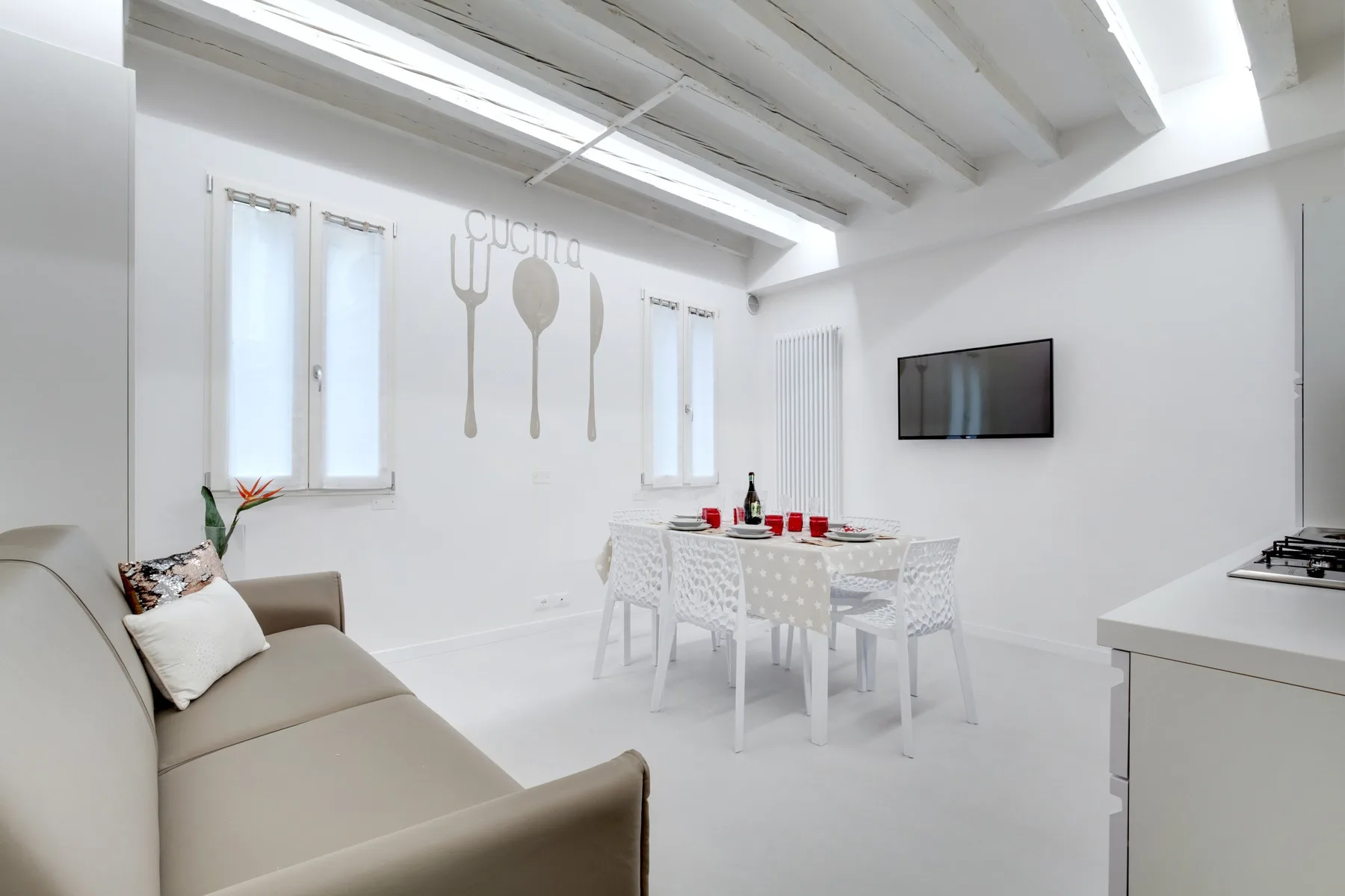 A bright dining area has a white table and chairs beside a wall-mounted television.