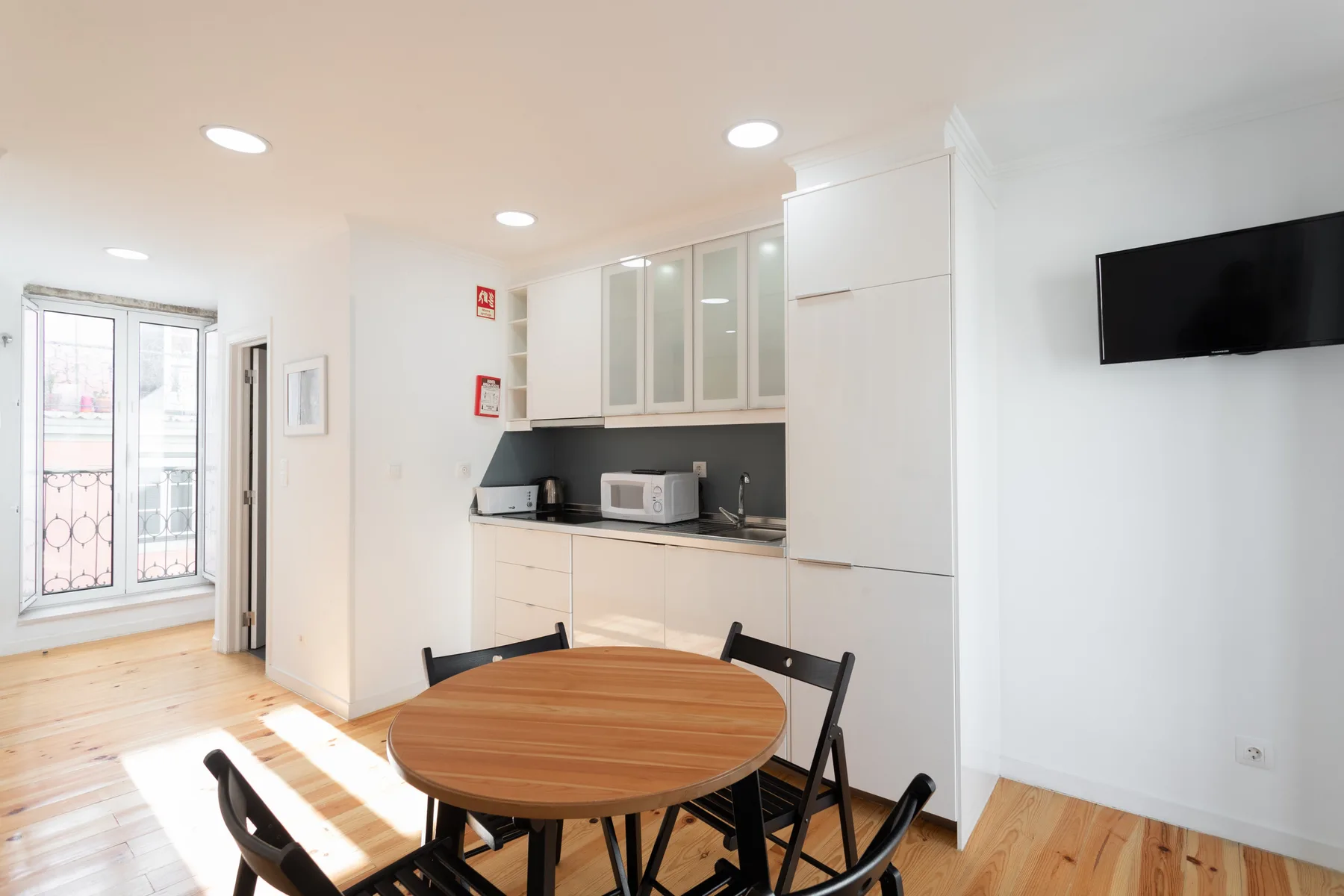 Una cocina y comedor luminosos con una mesa redonda de madera y un televisor en la pared.