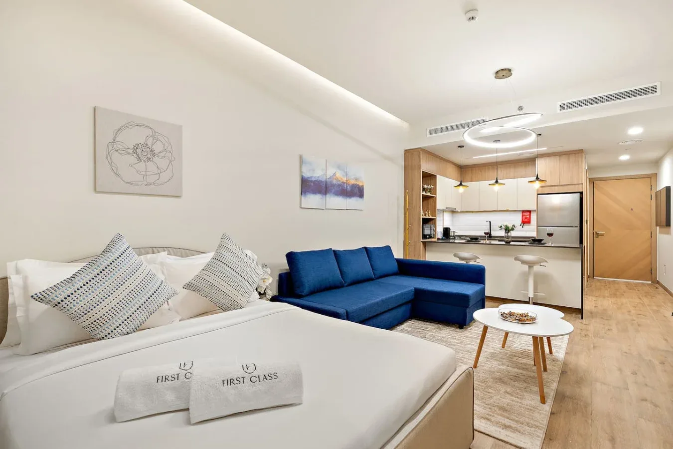 A modern studio living area with a bed, blue seating, and an open kitchen.