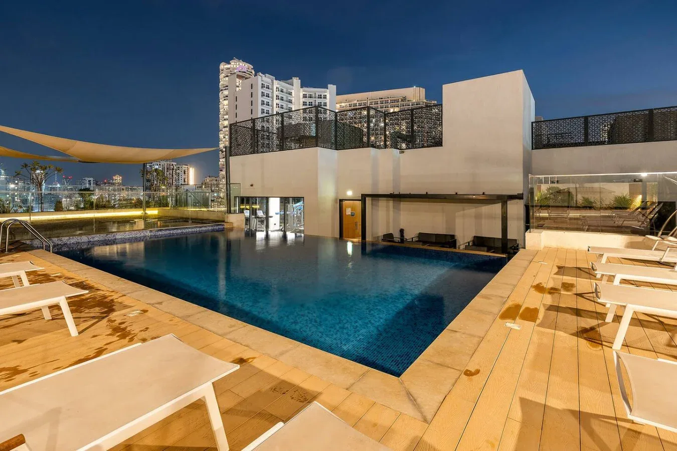 A rooftop swimming pool is surrounded by lounge chairs and city views at night.