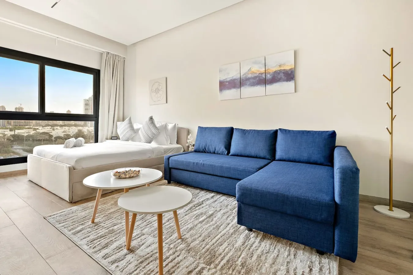 A bright living area combines blue seating, a white bed, and large windows.