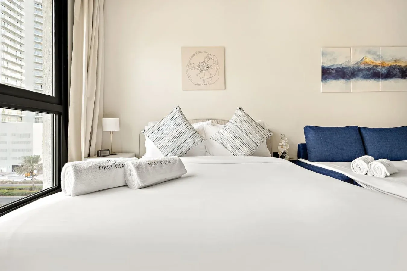 A bright bedroom with white bedding, decorative pillows, and a blue upholstered headboard.