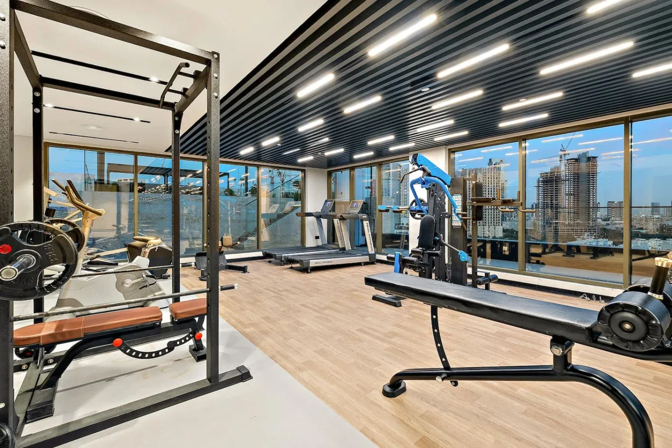 A gym with exercise machines and weight equipment beside large windows.