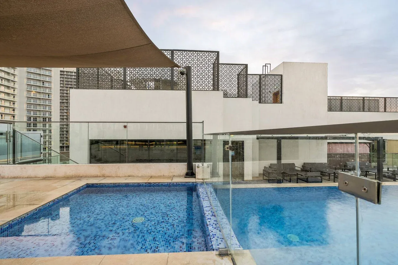 An outdoor swimming pool sits beside a modern white building and shaded seating.