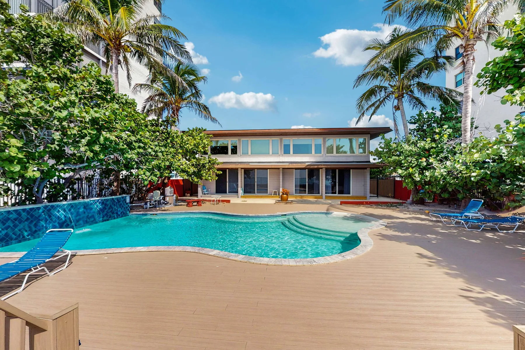 A swimming pool with curved steps sits beside a modern building and palm trees.