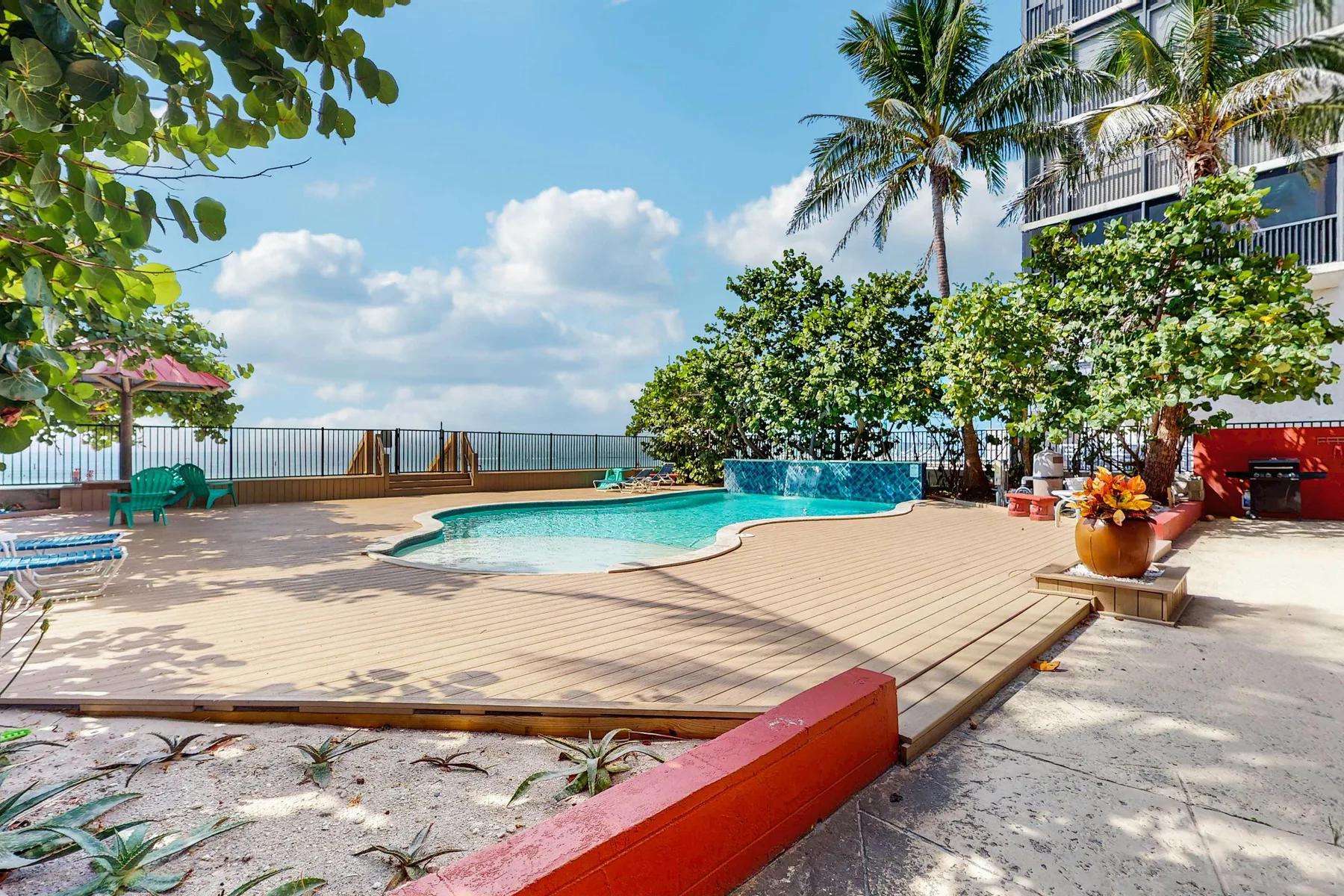 An outdoor pool and paved deck sit beside tropical plants and a railing.