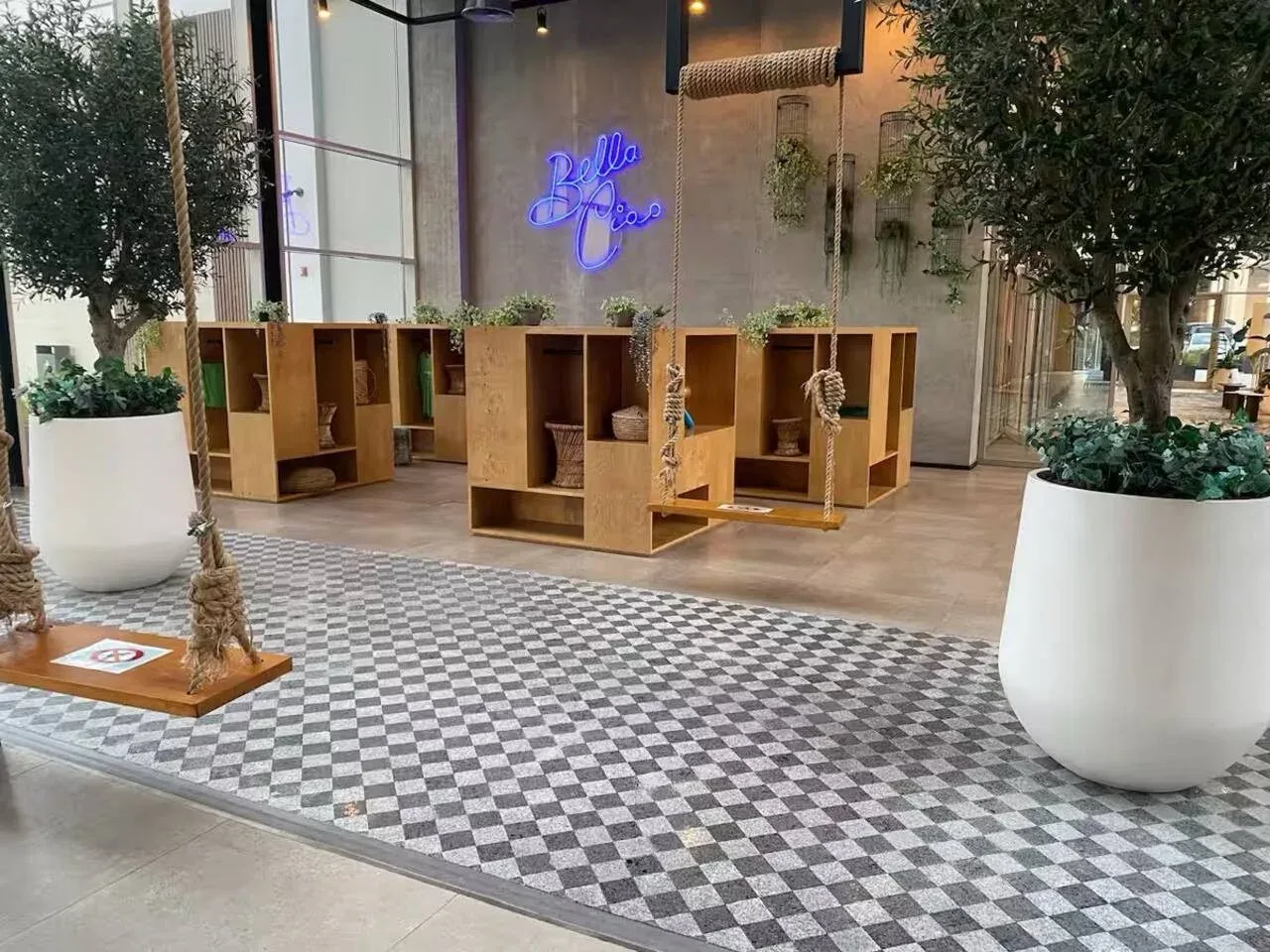An indoor lobby features wooden shelving, hanging swings, and large potted trees.
