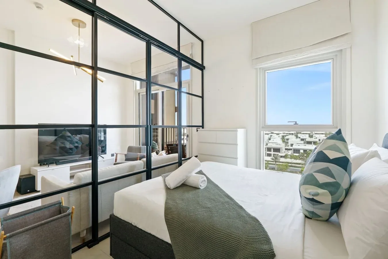 A bright bedroom with white bedding and a large window overlooking nearby buildings.