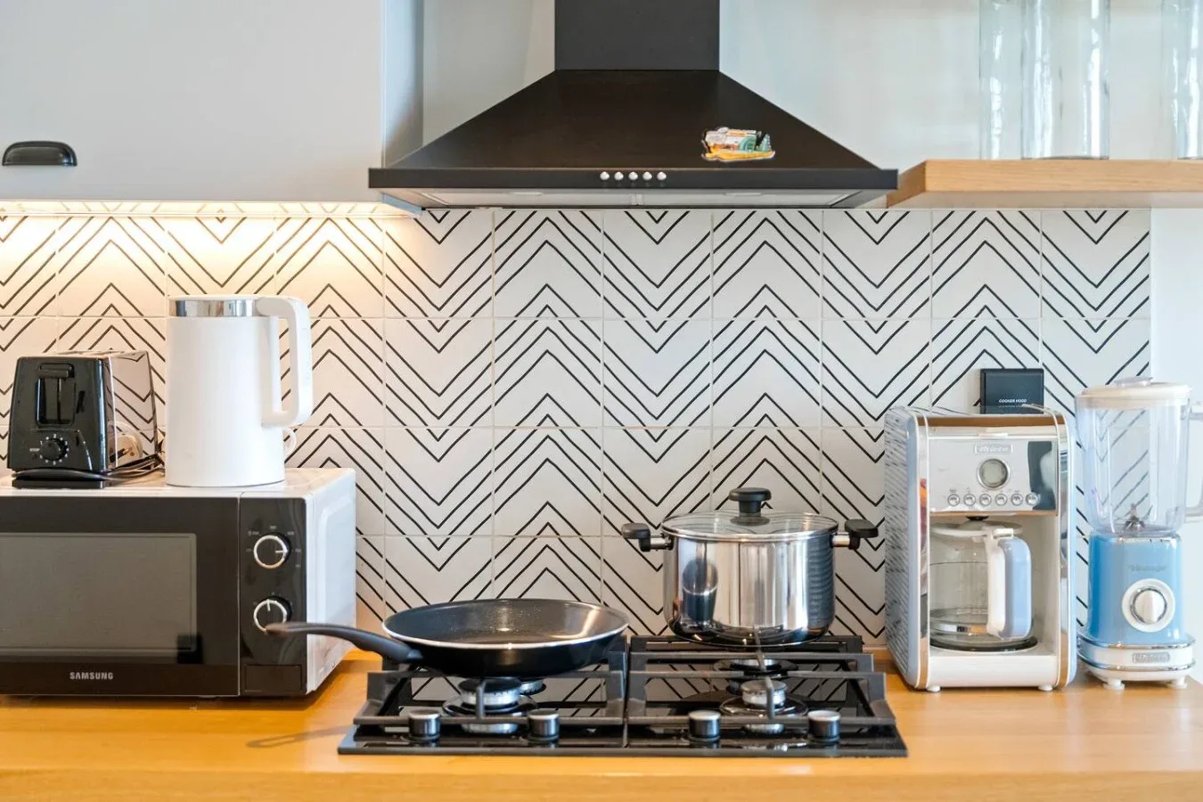 A kitchen counter with a stovetop, cookware, and small appliances against a patterned backsplash.