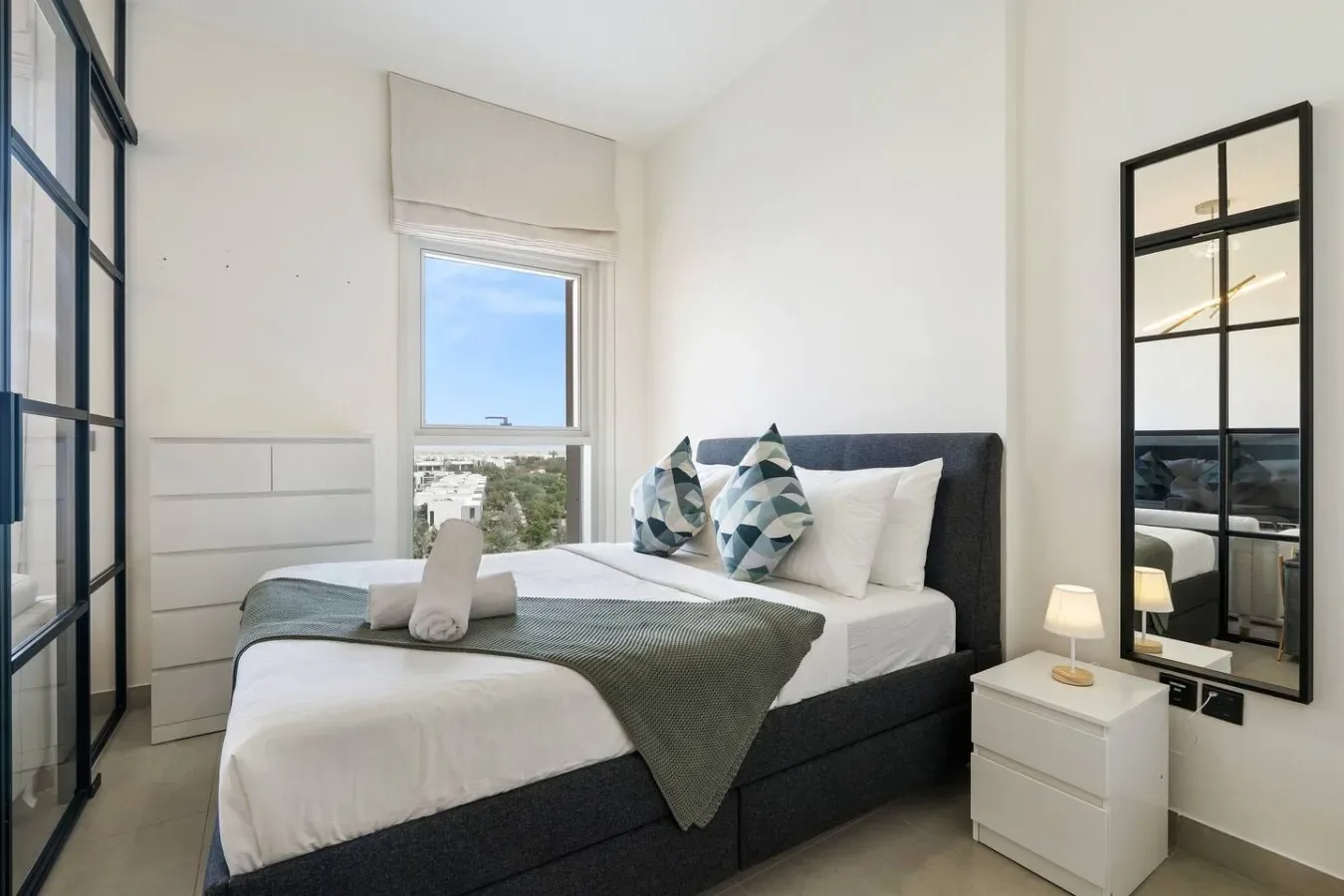 A bedroom with a neatly made bed beside a window and mirrored wardrobe.