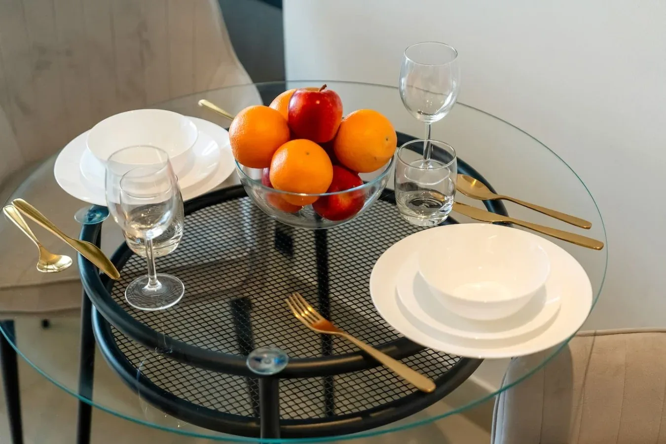 A dining table is set with plates, glasses, cutlery, and a bowl of fruit.
