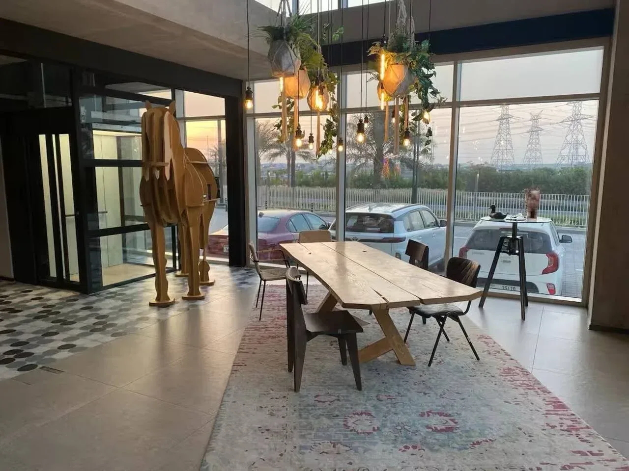 A dining area with a long table, chairs, and hanging lights beside large windows.