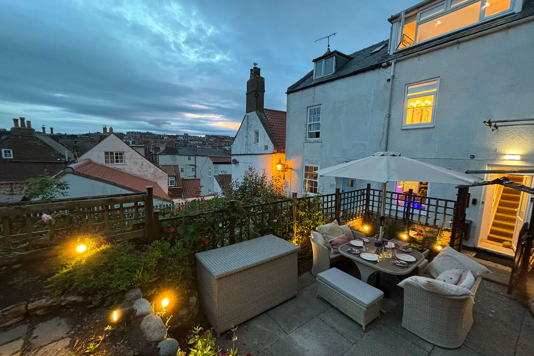 A rooftop terrace with outdoor seating, a table, and potted plants overlooks nearby buildings.