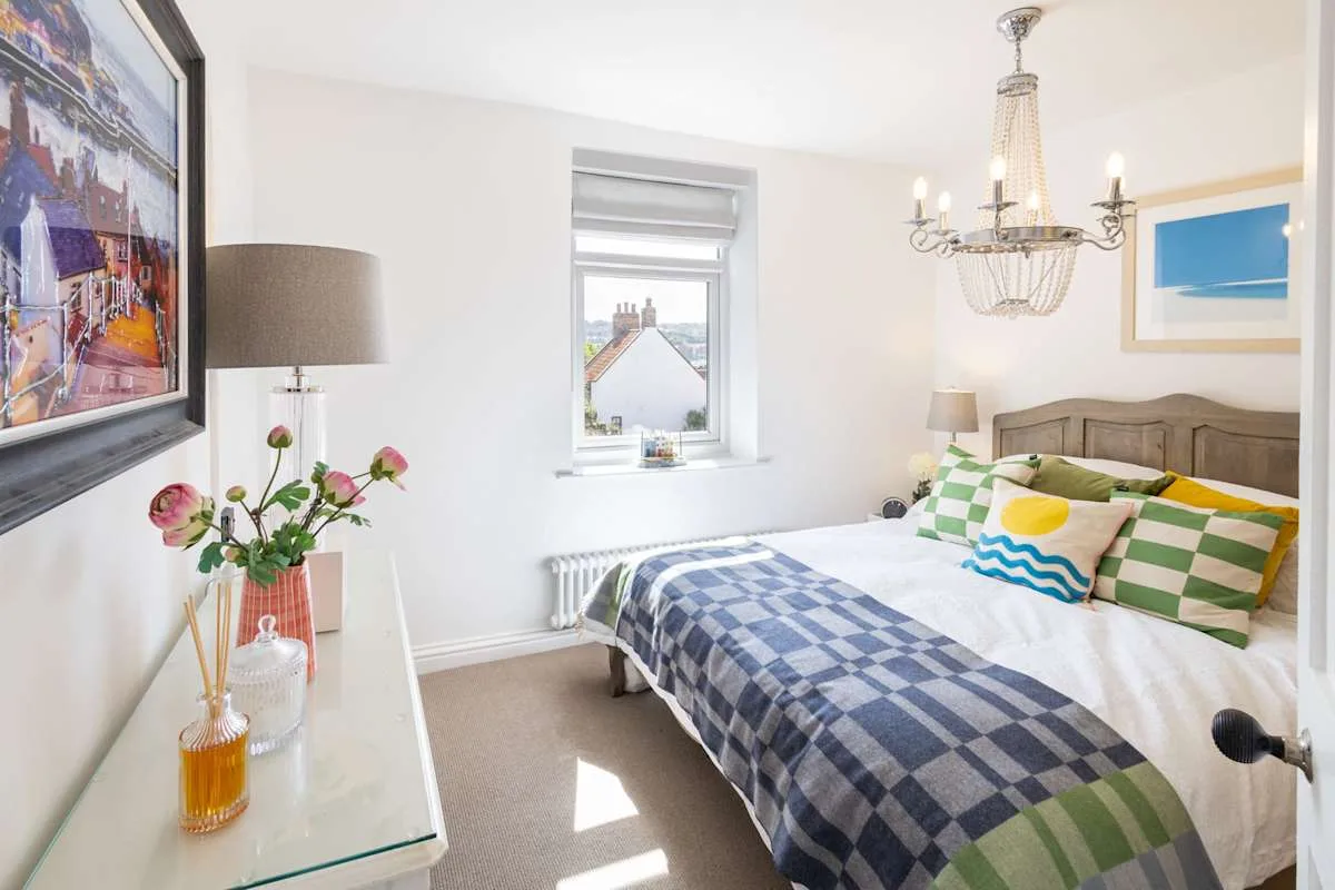 A bright bedroom with a bed beneath a chandelier and a window overlooking rooftops.