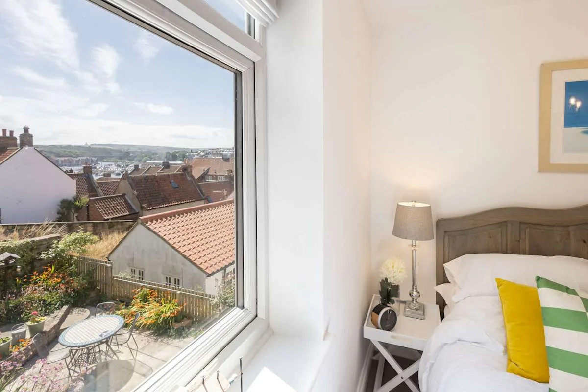 Bedroom with a bed beside a large window overlooking rooftops and a garden.