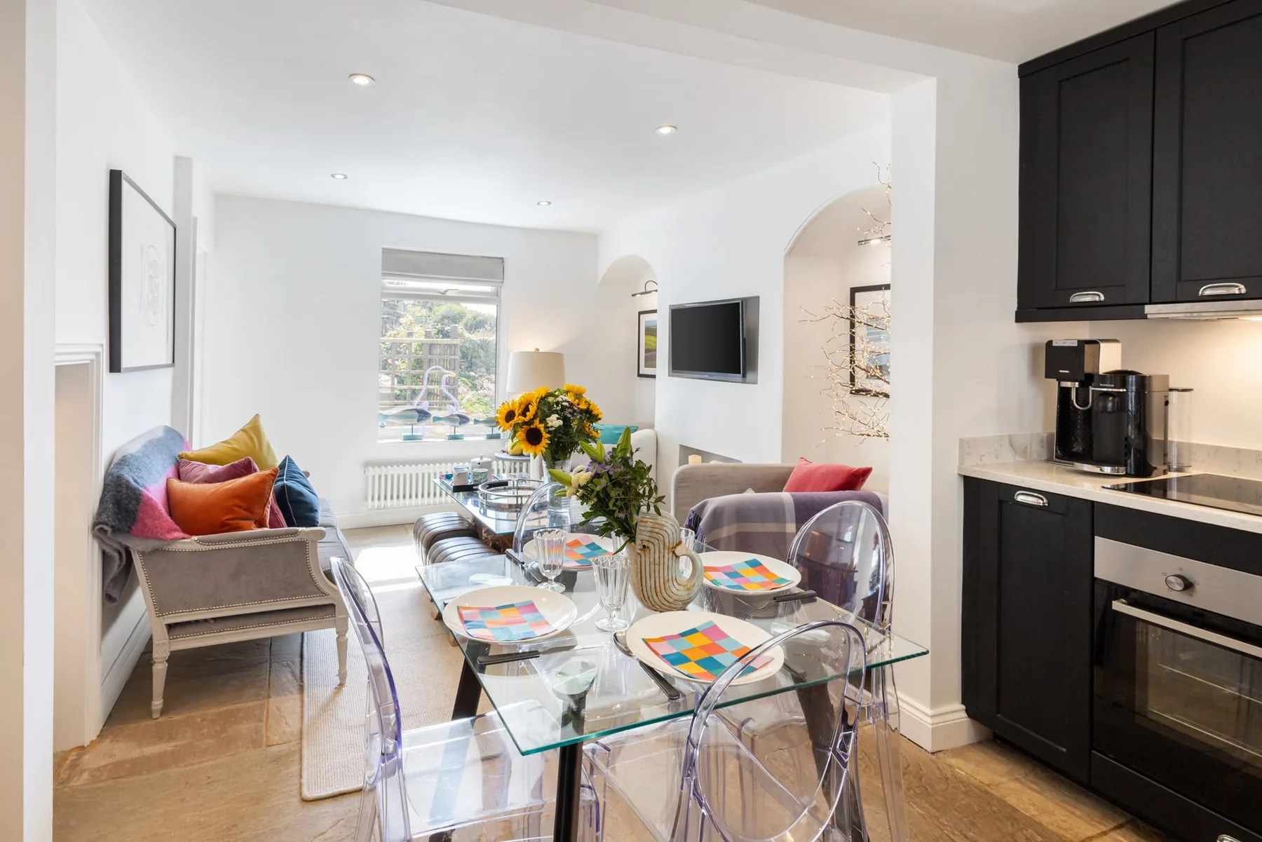 A bright dining and living area with a glass table and colorful seating.