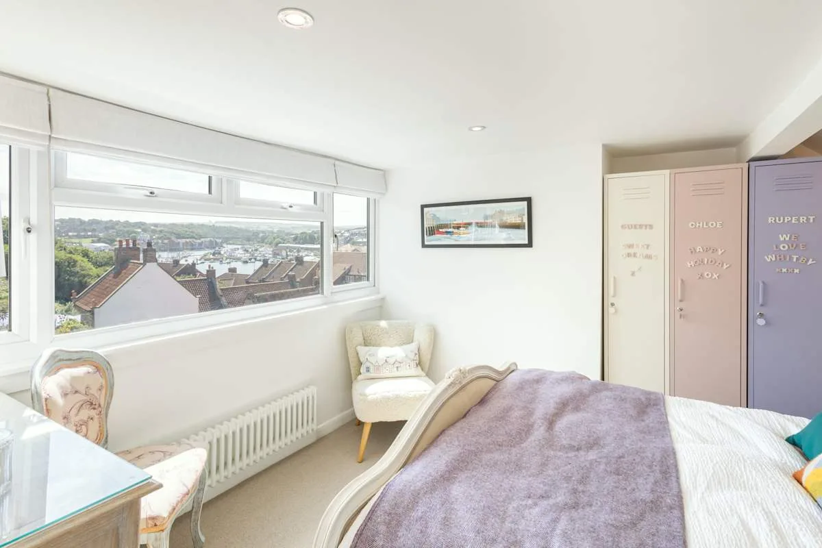 A bright bedroom with a bed, seating, and wide windows overlooking rooftops.